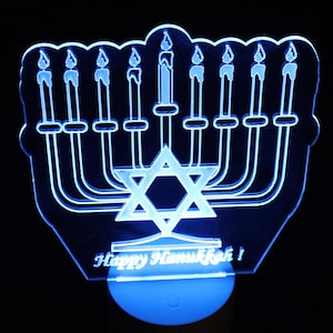 May include: A blue LED Hanukkah menorah night light. The menorah features nine candle holders, a Star of David, and the text "Happy Hanukkah!" The light is on a round base and glows blue against a black background.