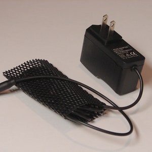 May include: Black AC adapter with a black power cord and a black mesh cable organizer. The adapter has a US plug and the text "AC/DC ADAPTER" printed on it.
