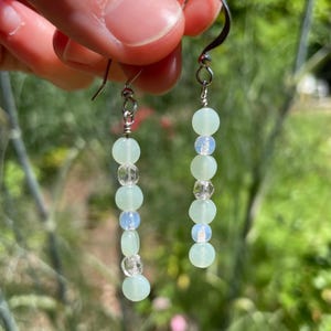 May include: A pair of dangle earrings featuring a vertical arrangement of alternating pale green round beads, clear faceted beads, and iridescent white beads. The beads are strung on silver-toned metal wire and attached to silver-toned metal hooks.