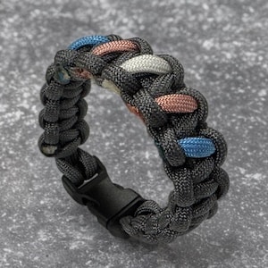 May include: A black paracord bracelet with a woven design featuring blue, white, and pink accents. The bracelet has a black plastic buckle closure.