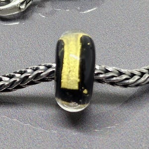 Op de afbeelding: Een zilveren armband met een visvormige sluiting en een zwart-gouden kraal. De armband heeft een geweven kettingontwerp. De kraal heeft een gouden streepontwerp. De sluiting is gedetailleerd met schubben.