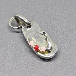 Könnte beinhalten: Ein silberfarbener Anhänger in Form einer Flip-Flop-Sandale. Die Sandale hat rosa und rote Sternakzente. Der Anhänger hat eine kleine Schlaufe zum Befestigen an einer Kette oder einem Armband. Der Hintergrund ist einfarbig grau.