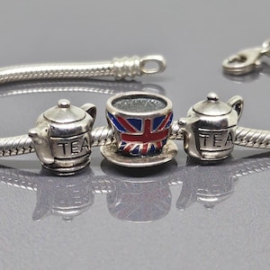 Könnte beinhalten: Ein silbernes Charm-Armband mit drei Anhängern. Zwei Anhänger sind Miniatur-Teekannen mit dem Wort "TEA". Der mittlere Anhänger ist eine Tasse und Untertasse mit dem Union Jack-Design. Das Armband liegt auf einer grauen Oberfläche.