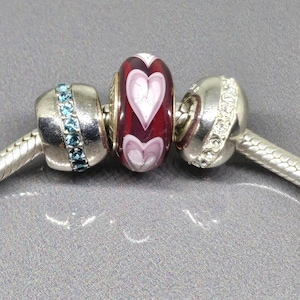 Peut inclure: Un bracelet en argent avec trois perles décoratives. La perle centrale est rouge foncé avec deux cœurs blancs. Les deux autres perles sont argentées avec des pierres bleues et transparentes.