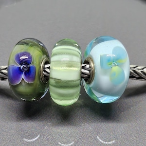 Peut inclure: Un bracelet en argent avec trois perles de verre. Les perles sont vertes, bleu clair et transparentes, avec des motifs floraux. Le bracelet a une chaîne tressée et un fermoir argenté.