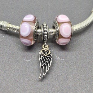 May include: A silver bracelet with two brown and pink polka-dot beads and a silver wing charm with a gold heart. The bracelet is on a gray surface.