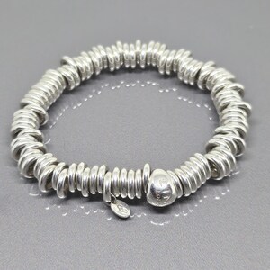 Puede incluir: Una pulsera de tono plateado compuesta por numerosos anillos metálicos circulares pequeños unidos en un bucle continuo. La pulsera presenta un único dije ligeramente más grande, liso y redondeado que cuelga de la cadena metálica.