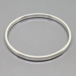 May include: A simple, silver-colored bangle bracelet. The round bracelet is made of a solid, shiny metal and has a smooth, flat surface. The bracelet is approximately 0.25 inches wide and has a minimalist design.