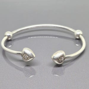 May include: A silver-toned open cuff bracelet. The bracelet features two heart-shaped ends, each adorned with small, sparkling embellishments. Two small spheres are on the bracelet's curved band. The bracelet is displayed on a gray surface.
