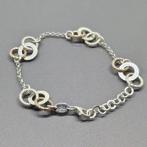 May include: A silver bracelet with a mix of textured and smooth circular links, interspersed with gold-toned accents. The bracelet has a lobster clasp closure and is displayed on a grey surface.