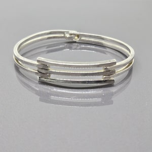 May include: Silver-toned bangle bracelet with a three-bar design. The bracelet features a hinged clasp and is composed of three parallel bands connected by rectangular bars. The jewelry is displayed on a reflective surface.