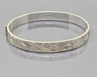 Könnte beinhalten: Ein silbernes, aufklappbares Armreif-Armband mit eingraviertem Blumenmuster. Das Armband hat einen Verschluss und eine polierte Oberfläche. Das Design zeigt detaillierte Blatt- und Wirbelmotive. Das Armband ist etwa 1,3 cm breit.