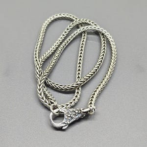 Op de afbeelding: Een zilverkleurige ketting met een gevlochten ontwerp. De ketting heeft een karabijnsluiting met een bloemenontwerp. De ketting wordt gepresenteerd op een grijze ondergrond.