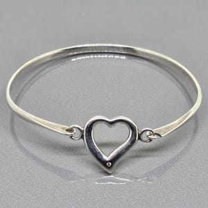 May include: A silver bangle bracelet with a heart-shaped centerpiece. The heart is open in the center and has a small, round accent at the bottom. The bracelet is made of a shiny, polished metal.