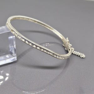 May include: A silver-toned hinged bracelet with a row of small, clear crystals set along the top edge. The bracelet has a simple, elegant design and a small chain attached to the clasp. The background is a solid gray.