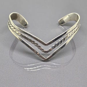 May include: A silver bangle bracelet with a V-shaped design. The bracelet has a braided pattern on the top and bottom edges.