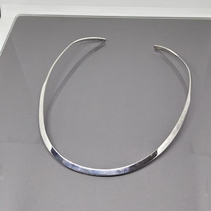 Może przedstawiać: Srebrny naszyjnik typu choker o płaskiej, zakrzywionej konstrukcji. Naszyjnik jest otwarty z przodu i spoczywa na szarej powierzchni. Metal ma polerowane, odblaskowe wykończenie, tworząc elegancki i nowoczesny wygląd.