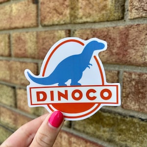May include: A sticker featuring a blue dinosaur silhouette inside a white circle with an orange border. The word "DINOCO" is displayed in white letters on an orange rectangle below the dinosaur image. The sticker is held against a brick wall.