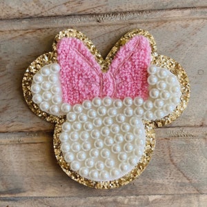 May include: A decorative Minnie Mouse-shaped embellishment. It features a pink bow, white pearl accents, and a gold glitter outline. Likely for crafts or accessories.