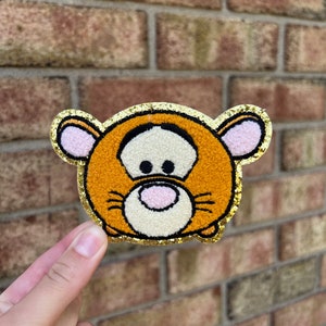 May include: A plush orange and brown Tigger character with a gold glitter outline. The character is in a round, flat shape, resembling a popular Japanese character.