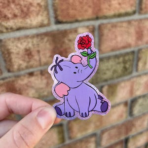 May include: A purple glitter sticker of a cartoon elephant holding a red rose. The elephant is sitting down and has a pink ear.