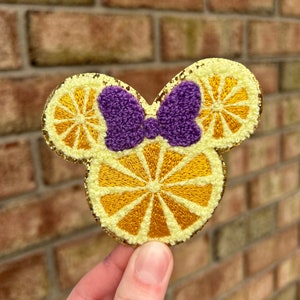 May include: A yellow and gold embroidered lemon-shaped Mickey Mouse ears with a purple bow.
