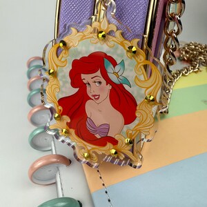 May include: A clear acrylic charm featuring a cartoon image of Ariel from The Little Mermaid, set within a gold-colored ornate frame. The charm is adorned with small gold-colored embellishments and hangs from a gold chain. The background includes a purple purse and a pastel-colored notebook.