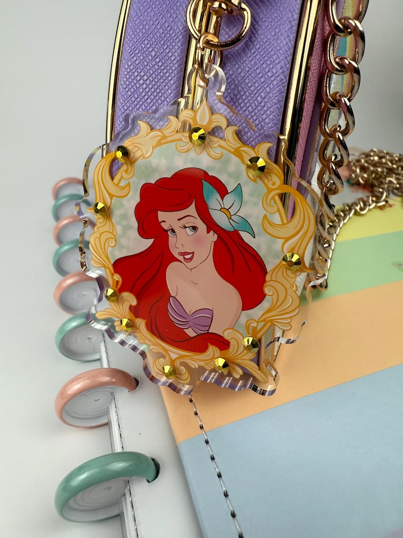 May include: A clear acrylic charm featuring a cartoon image of Ariel from The Little Mermaid, set within a gold-colored ornate frame. The charm is adorned with small gold-colored embellishments and hangs from a gold chain. The background includes a purple purse and a pastel-colored notebook.