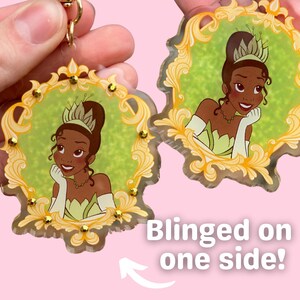 May include: Two princess-themed keychains. Each keychain features a princess illustration within a gold-colored ornate frame, accented with gold-colored embellishments. The text "Blinged on one side!" is visible. The keychains are held against a pink background.