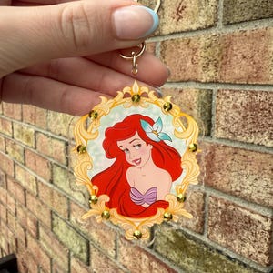May include: A clear acrylic keychain featuring a portrait of Ariel from The Little Mermaid. The image is framed in a gold-colored ornate border with gold-colored embellishments. The keychain has a gold-colored ring and clasp.