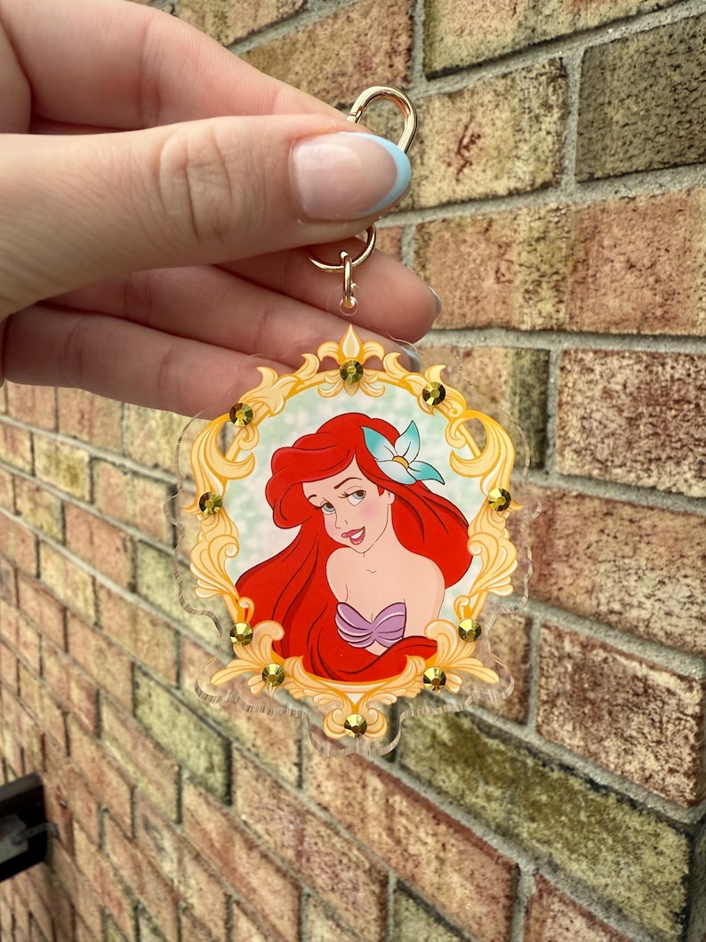 May include: A clear acrylic keychain featuring a portrait of Ariel from The Little Mermaid. The image is framed in a gold-colored ornate border with gold-colored embellishments. The keychain has a gold-colored ring and clasp.