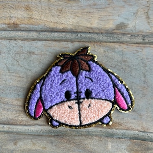 May include: Purple and pink embroidered plush character with a brown head and gold glitter trim.