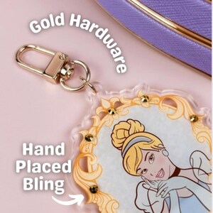 May include: A clear acrylic keychain featuring a Cinderella illustration with gold accents and hand-placed bling. The keychain has a gold-colored clasp and is set against a pink background. The text "Gold Hardware" and "Hand Placed Bling" are visible.