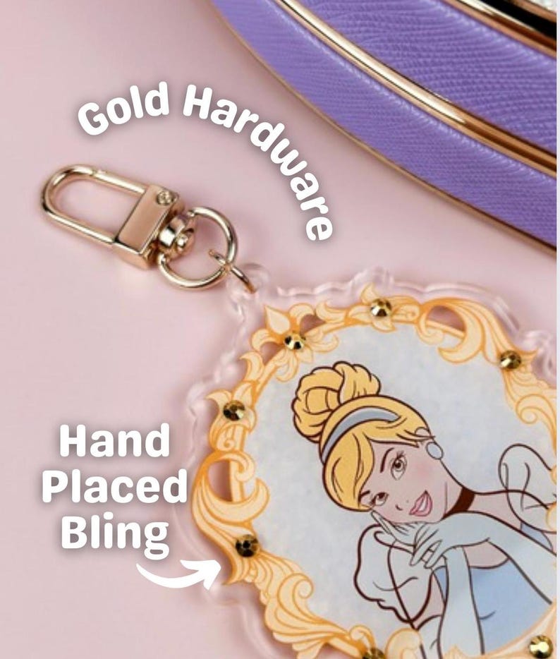 May include: A clear acrylic keychain featuring a Cinderella illustration with gold accents and hand-placed bling. The keychain has a gold-colored clasp and is set against a pink background. The text "Gold Hardware" and "Hand Placed Bling" are visible.