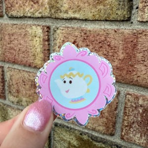 May include: A pink sticker with a scalloped edge and a glittery border. The sticker features a cartoon teapot character inside a light blue circle. The teapot has a smiling face and yellow accents.