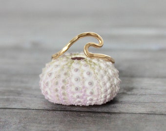 Pode incluir: Um anel de onda de ouro, uma peça de joalheria simples e delicada, é exibido em um ouriço-do-mar branco. O anel é feito de um único pedaço de fio que é dobrado na forma de uma onda.