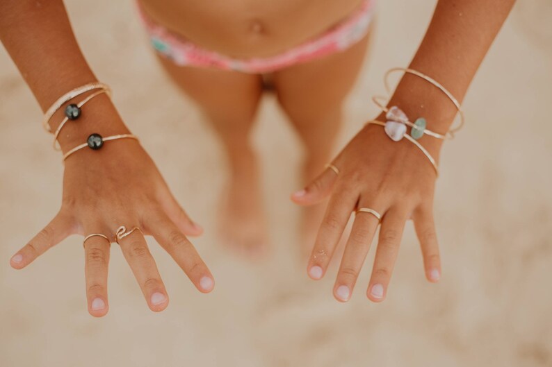 Pode incluir: Close-up das m&atilde;os de uma pessoa usando an&eacute;is e pulseiras de ouro. As pulseiras t&ecirc;m p&eacute;rolas pretas e pequenas pedras. As unhas da pessoa est&atilde;o pintadas de uma cor rosa claro.