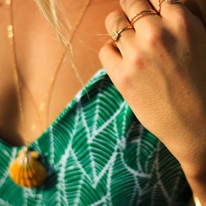 Pode incluir: Uma mulher vestindo um top estampado verde e branco com um colar de ouro e um pingente de concha. Ela est&aacute; usando v&aacute;rios an&eacute;is de ouro nos dedos.