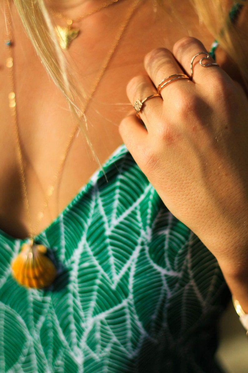Pode incluir: Uma mulher vestindo um top estampado verde e branco com um colar de ouro e um pingente de concha. Ela est&aacute; usando v&aacute;rios an&eacute;is de ouro nos dedos.