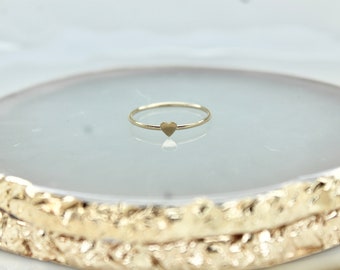 Pode incluir: Um anel de ouro simples com um pequeno design em forma de coração. O anel está sobre uma superfície texturizada branca e dourada.