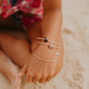 Pode incluir: Uma pessoa usando tr&ecirc;s pulseiras de ouro no pulso. As pulseiras s&atilde;o adornadas com uma p&eacute;rola preta e uma pequena concha.