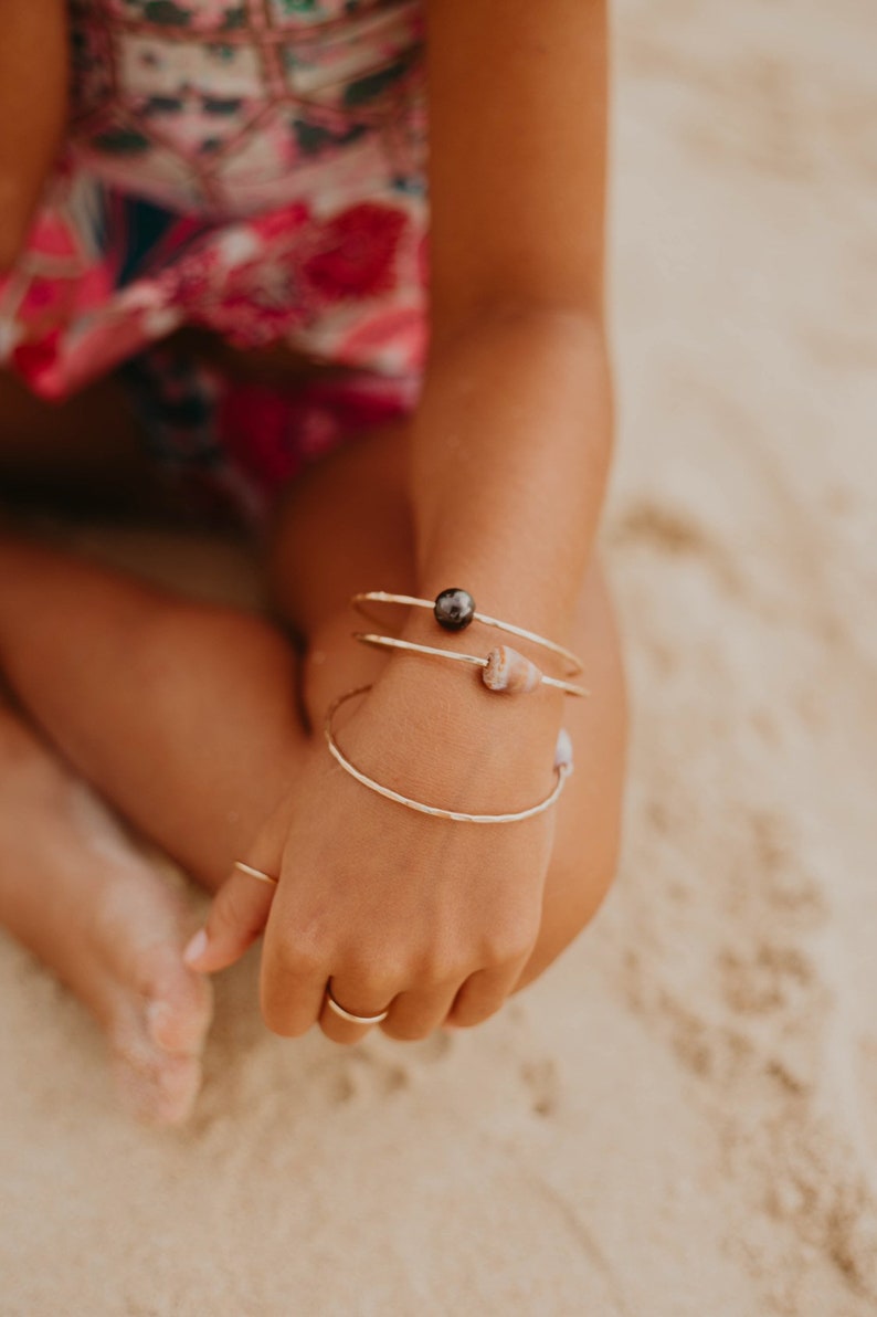 Pode incluir: Uma pessoa usando tr&ecirc;s pulseiras de ouro no pulso. As pulseiras s&atilde;o adornadas com uma p&eacute;rola preta e uma pequena concha.