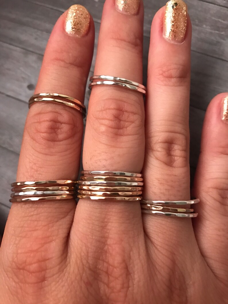 Pode incluir: Um conjunto de cinco an&eacute;is de metal martelado em tons de prata e ouro. Os an&eacute;is est&atilde;o empilhados em um dedo.