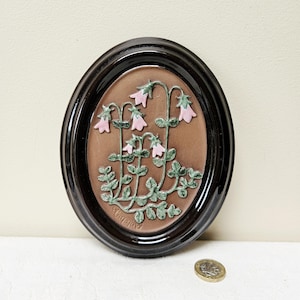 Può includere: Opera d'arte murale ovale in ceramica con rilievo di fiori rosa a forma di campana e foglie verdi su sfondo marrone. Incorniciata in una cornice ovale marrone scuro lucido. L'opera misura circa 15 cm di altezza.