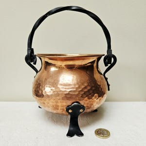 May include: A copper cauldron with a hammered finish, featuring a black wrought iron handle and legs. The cauldron has a rounded shape. A coin is next to the cauldron.