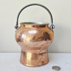 May include: A hammered copper pot with a rounded body and a flared base, featuring a dark metal swing handle attached to two side loops. The interior is lined with a silver-coloured metal. A small circular coin is placed next to the pot for scale.