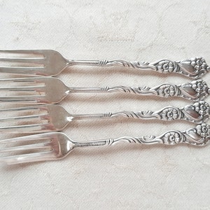 Peut inclure: Quatre fourchettes en argent avec des détails floraux ornementaux sur les manches. Les dents sont espacées uniformément. Les fourchettes sont disposées horizontalement sur une surface blanche et texturée, suggérant un cadre de restauration formel.