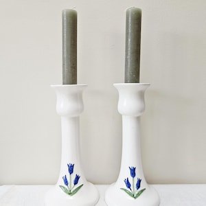 May include: Two white ceramic candle holders with a flared base and top, each featuring a hand-painted blue tulip design with green leaves on the front. Each holder contains a tall, slender, muted sage green taper candle standing upright.