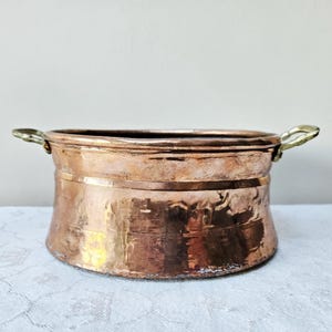 Könnte beinhalten: Ein gehämmertes Kupfergefäß mit Messinggriffen. Der Topf hat einen breiten, abgerundeten Körper und einen leicht ausgestellten Rand. Das Kupfer hat eine warme, rötlich-braune Farbe mit dunkleren Patina-Bereichen. Die Griffe sind gebogen und an den Seiten des Topfes befestigt.
