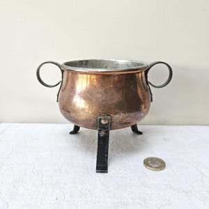 Puede incluir: Una olla de cobre con cuerpo redondeado, interior plateado y dos asas laterales de metal oscuro. La olla se apoya sobre tres patas de metal oscuro fijadas con remaches. Una moneda está colocada al lado para servir de referencia de escala.